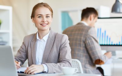 Happy businesswoman making notes in working diary with her colleague on background